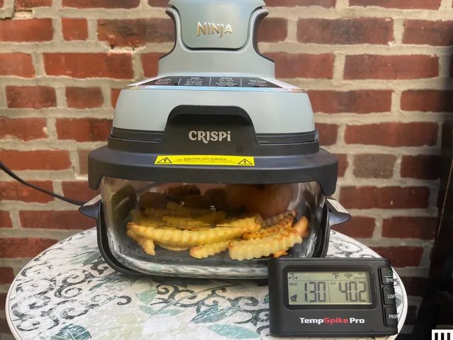 Closeup of the Ninja Crispi, a portable airfryer comprised of a clear dish on the bottom and a two-tier funnel-shaped device attached on top, sitting on a small table outdoors with a digital thermometer in front