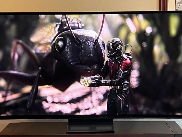 Front view of Samsung S90D Q.D.-O.L.E.D., a large screen tv showing a movie scene of a miniature human in a special superhero suit standing beside a bug.